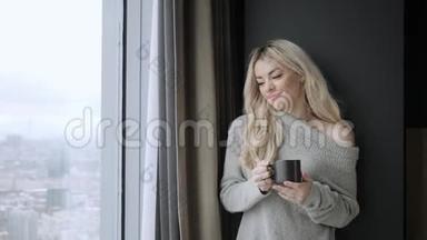 美丽的金发女人拿着陶瓷杯的茶或咖啡享受。 在床上喝茶。 看着窗户喝茶